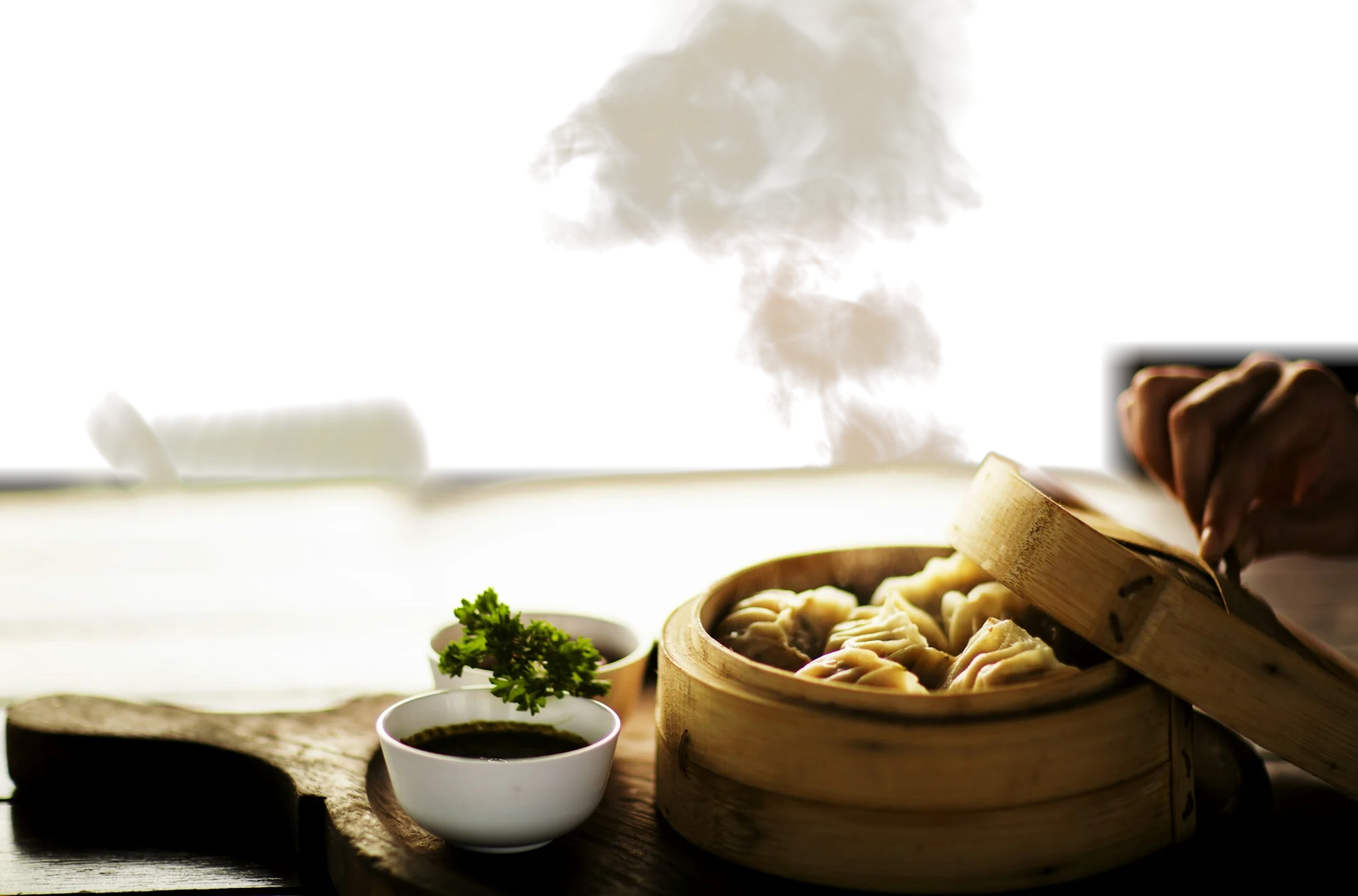 Steam rising from a bamboo basket of dumplings beside a small bowl of dipping sauce on a dark wooden board