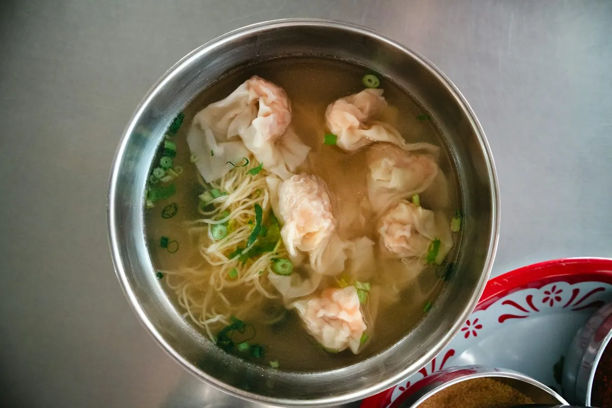 A metal bowl of thin egg noodles with shrimp wontons and spring onion in clear broth